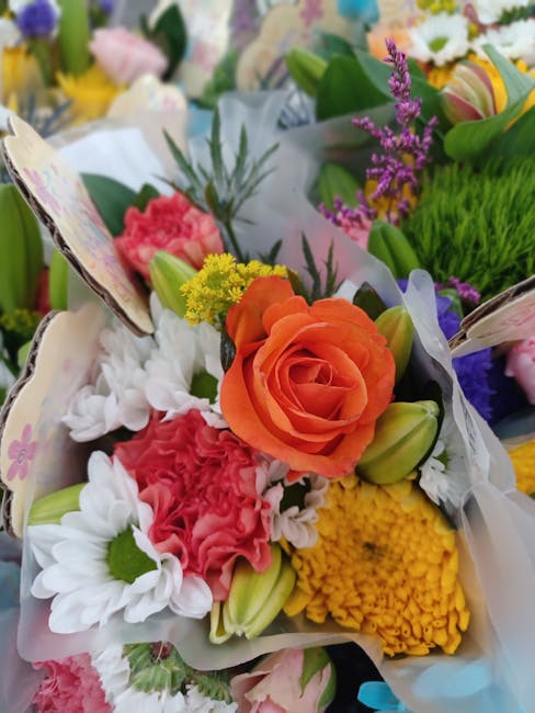 A vibrant floral bouquet arranged with a large, peach-colored rose at the center, surrounded by white daisies with green centers, pink carnations, yellow button chrysanthemums, and sprigs of purple la