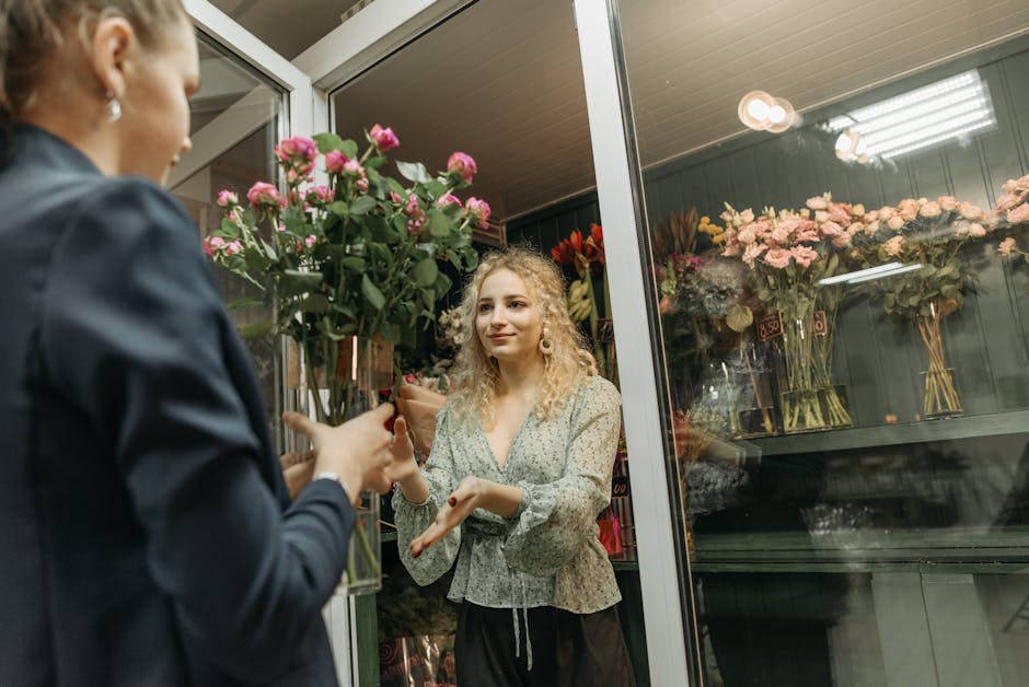 A customer at flowerdeliverystroudgreen.co.uk is inside a floral shop, engaging in a transaction with a florist who is handing over a bouquet of fresh pink roses with green foliage. The bouquet is wra