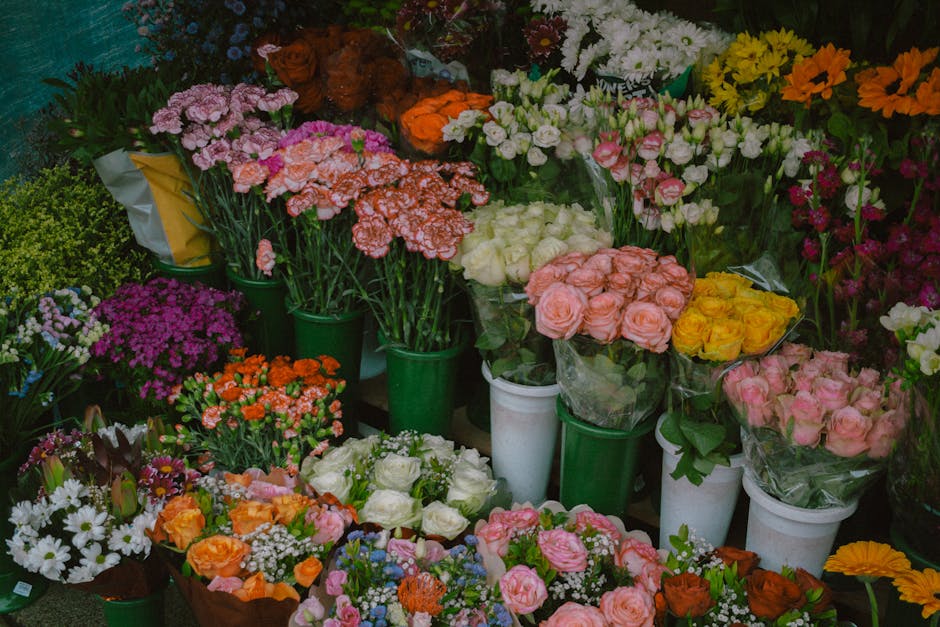 A vibrant display of fresh flowers arranged in various bouquets and potted plants, featuring roses in soft pink, creamy white, and bright yellow, alongside clusters of purple, orange, and white daisie