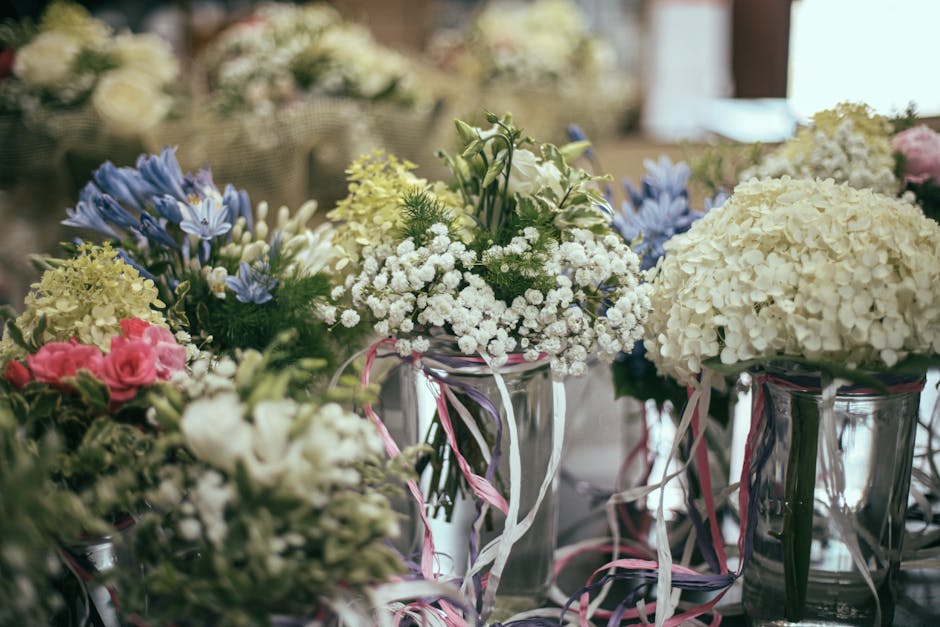 A collection of floral arrangements featuring white hydrangeas, pink roses, and blue delphiniums, displayed in clear glass vases decorated with pink and white ribbon accents. The flowers are fresh, wi