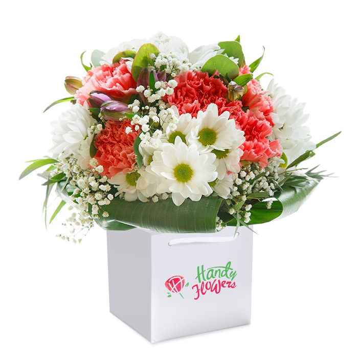 A beautifully composed bouquet sits upright in its pristine, square-edged white gift bag, set against a backdrop of pure white that lets every colour and texture speak for itself. At the heart of the arrangement are coral-pink carnations, their generous, ruffled petals lending depth and a satisfying richness to the ensemble. Bright white daisies stand beside them, each with a vibrant lime-green centre, their presence both striking and gently cheerful-like glimpsing wildflowers while strolling through Stroud Green's Woodberry Down. Subtle wisps of baby's breath interlace throughout, imparting an airy, feathery texture that brings lightness and lift. Scattered among the main blooms, unopened buds touched with hints of purple and pink pique curiosity, promising longevity and continued blossoming. These blooms are embraced by a collar of broad, glossy green leaves, which provide natural structure and a lively backdrop, framing the rounded shape in a way only a practiced Stroud Green florist might arrange. Finished with sturdy ribbon handles, the understated white gift bag makes the bouquet ready for easy delivery-perhaps to a home on Crouch Hill or for a special occasion in a local café. Each element of the bouquet radiates freshness, artistry, and thoughtful presentation, perfectly suited for birthdays, anniversaries, or gestures of affection, and reflecting the warm, creative heart of the Stroud Green community.